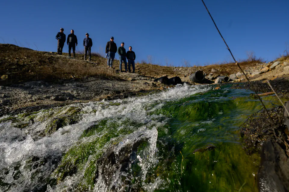 Waste water rushes over rocks covered in green organic material while a team of five people stand in the background.